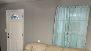 A beige leather sofa in a room with wooden flooring, a white door with a decorative glass panel, and a light blue curtain. - Entire Home  4-bedroom house in LakeWhitney Estates with WiFi, AC. Book now! (Whitney)