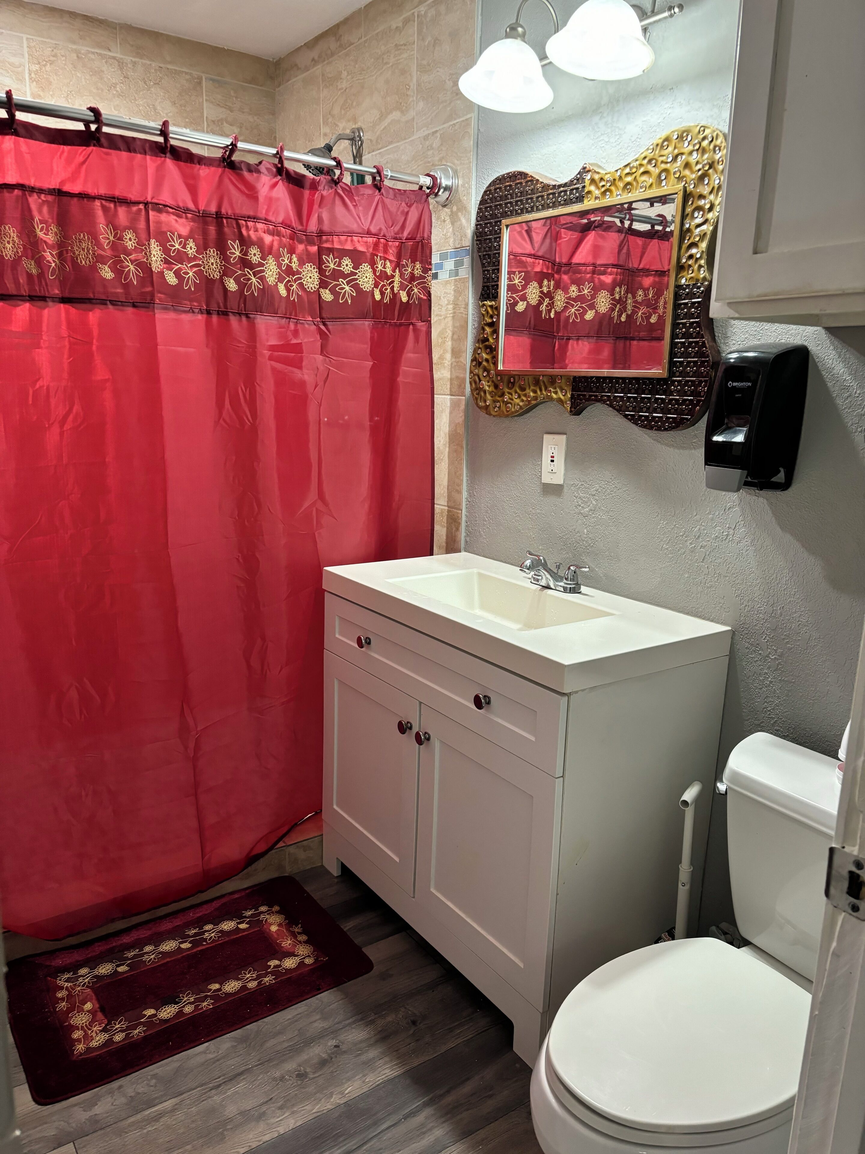 Une salle de bain avec un lavabo blanc, des toilettes, un rideau de douche rouge et un miroir doté d’un cadre décoratif.