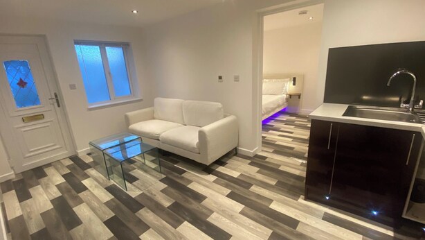A modern living room with a white sofa, a glass coffee table, and a kitchen area with a black countertop and sink. - Stunning Truro 1-bed - Sleeps 2 & Parking (Truro)