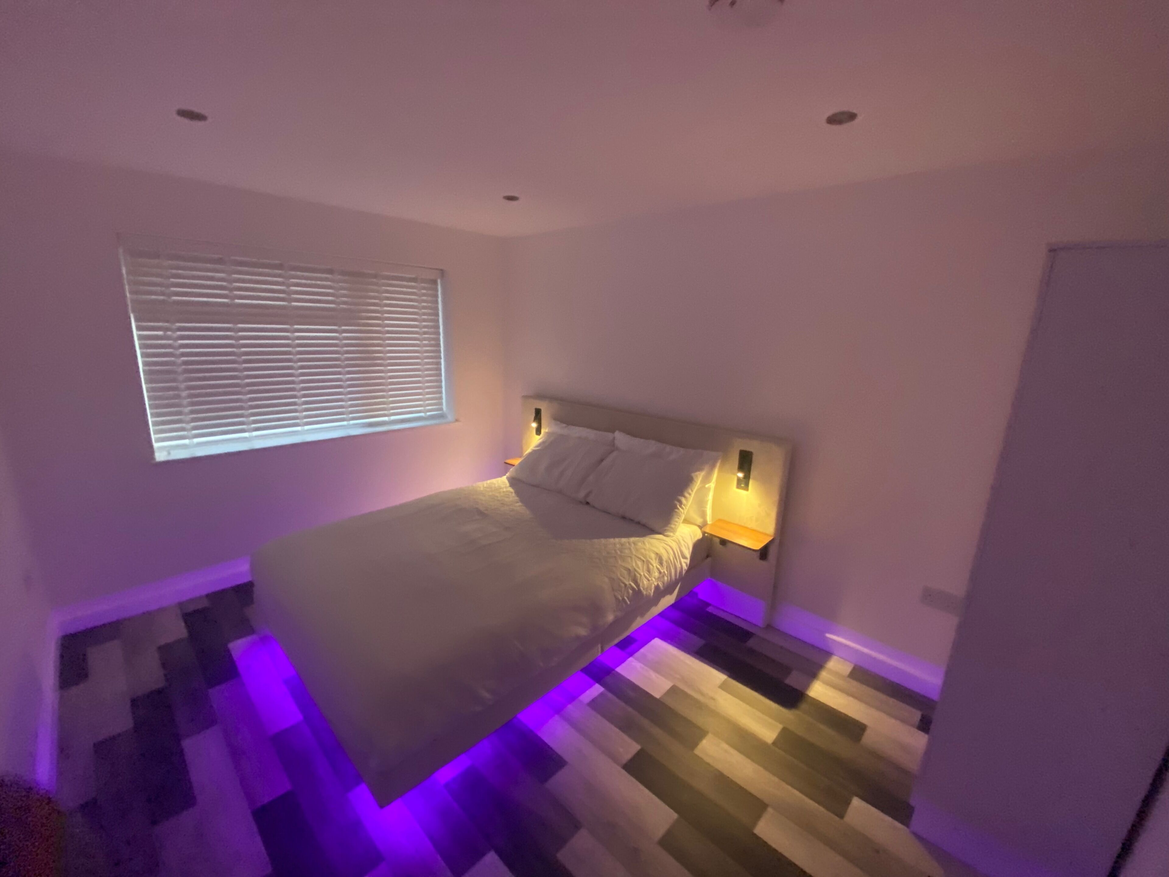 A bedroom with a bed, a window with blinds, a nightstand with a lamp, and a checkered floor.