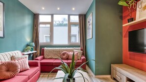 Sala de estar con sofá rojo, sofá beige y mueble de televisión de madera. Hay un ventanal con cortinas, una planta en maceta y un cuadro en la pared.