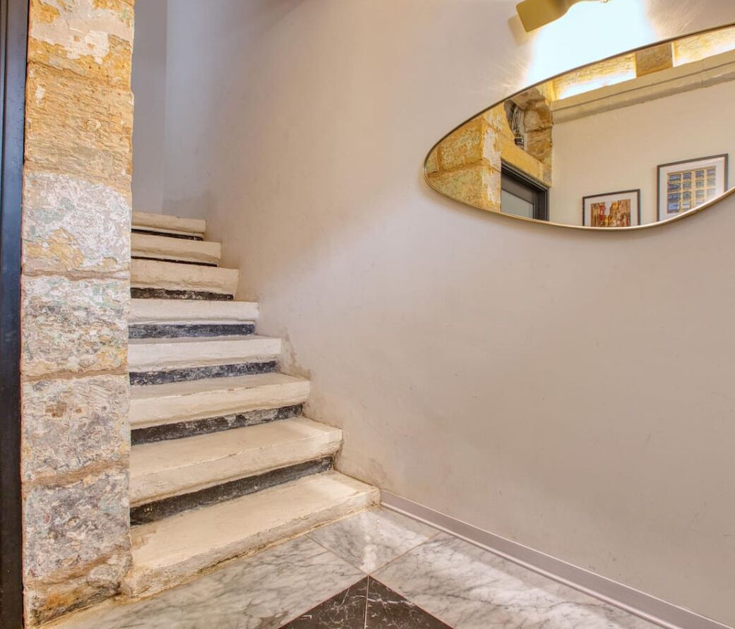 Una escalera de mármol con un muro de piedra en un lado, que conduce a un cuarto con un espejo grande que refleja un cuadro enmarcado.