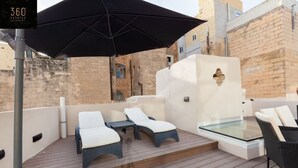 A rooftop terrace with lounge chairs and a glass table, surrounded by stone walls and a large black umbrella.
