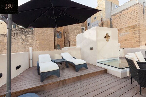 A rooftop terrace with lounge chairs and a glass table, surrounded by stone walls and a large black umbrella.
