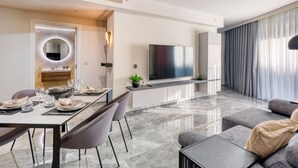 A modern living room with a grey sofa, a dining area with a table set for four, a TV mounted on the wall, and a large window with curtains. - The Official Mercury Suites: High end suite & WIFI (St. Julian`s)