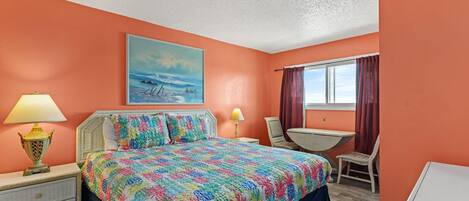 A bedroom with an orange wall, a bed with a colorful floral bedspread, two bedside lamps, a window with curtains, and a small table with a chair.