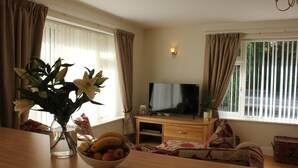 A living room with a TV, a wooden table with a bowl of fruit and a vase of flowers, and a sofa. - Quaint 1-bedroom house in Cheltenham (Cheltenham)