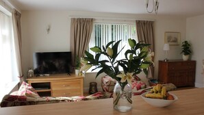 A living room with a wooden table, a vase with flowers, a television, and a window with curtains. - Quaint 1-bedroom house in Cheltenham (Cheltenham)