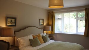 A neatly made bed with a green blanket, two bedside lamps, and a window with curtains. - Quaint 1-bedroom house in Cheltenham (Cheltenham)