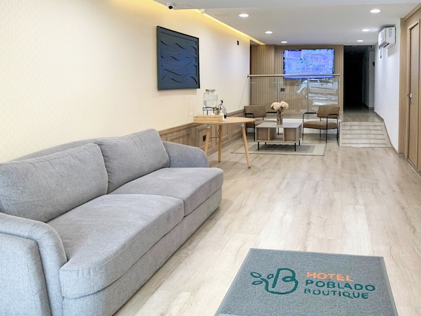 Un hall d’hôtel avec un canapé gris, une table en bois entourée de chaises, un écran de télévision et un paillasson portant le nom de l’hôtel.