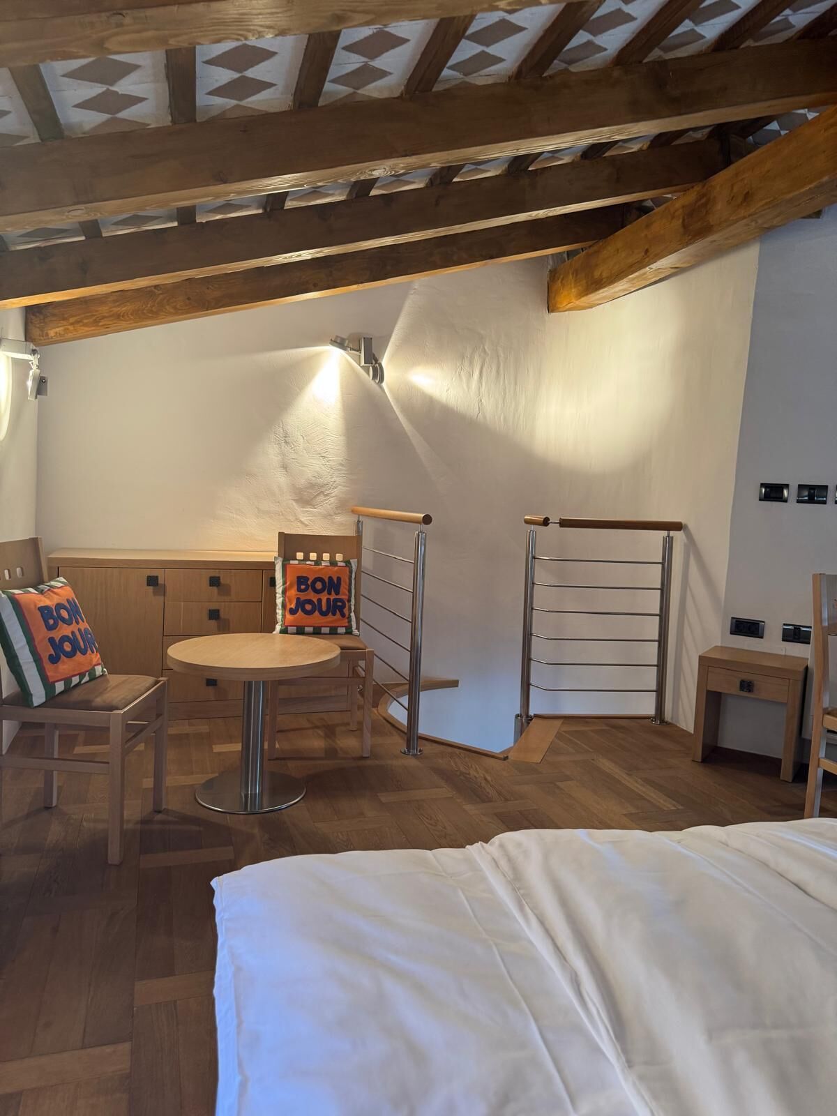 A cozy room with a bed, a wooden bench, a small table, and a chair. There's a 'BON JOUR' sign on the bench.