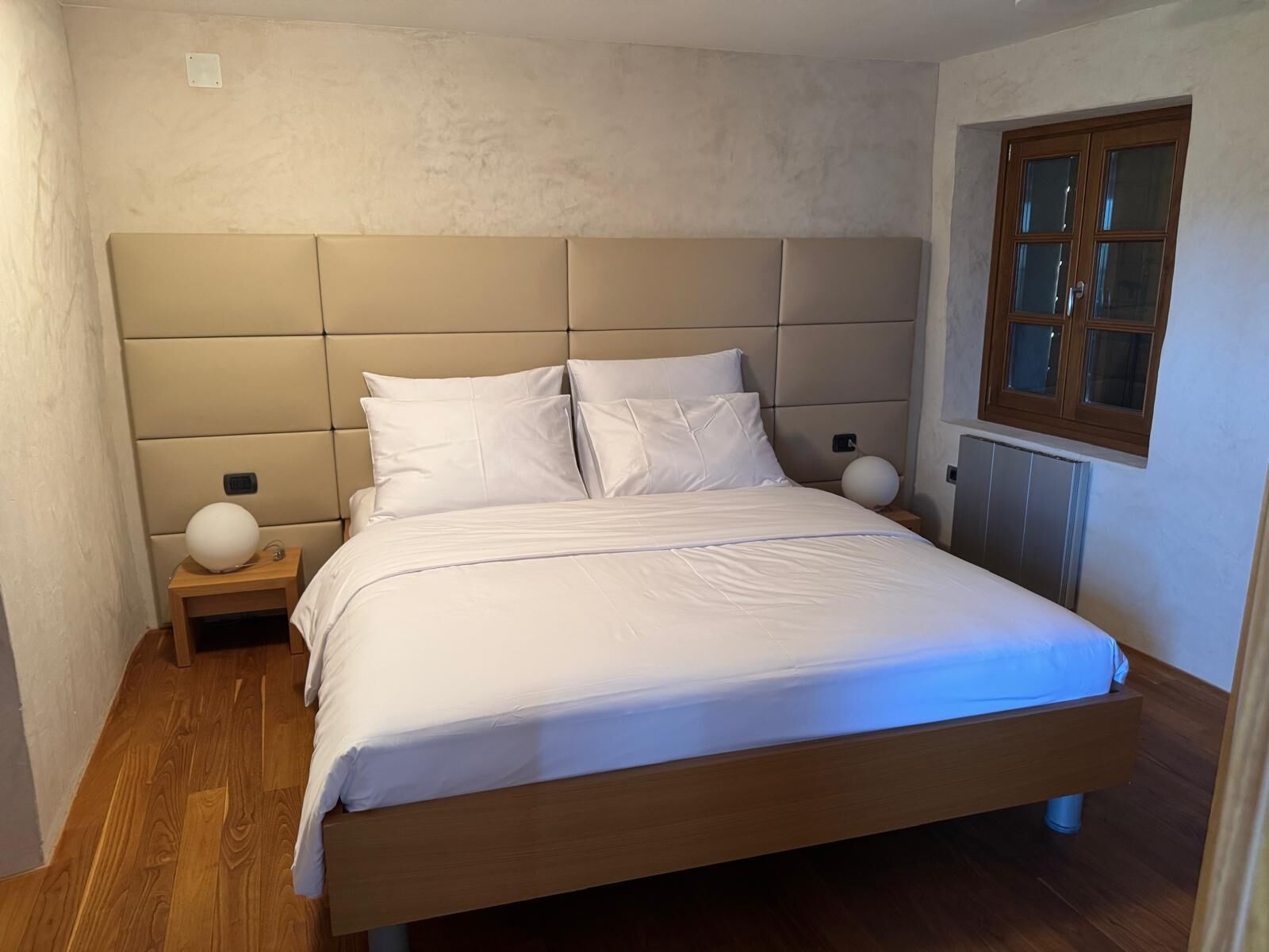 A bedroom with a large bed, a wooden headboard, a nightstand with a lamp, and a window with wooden shutters.