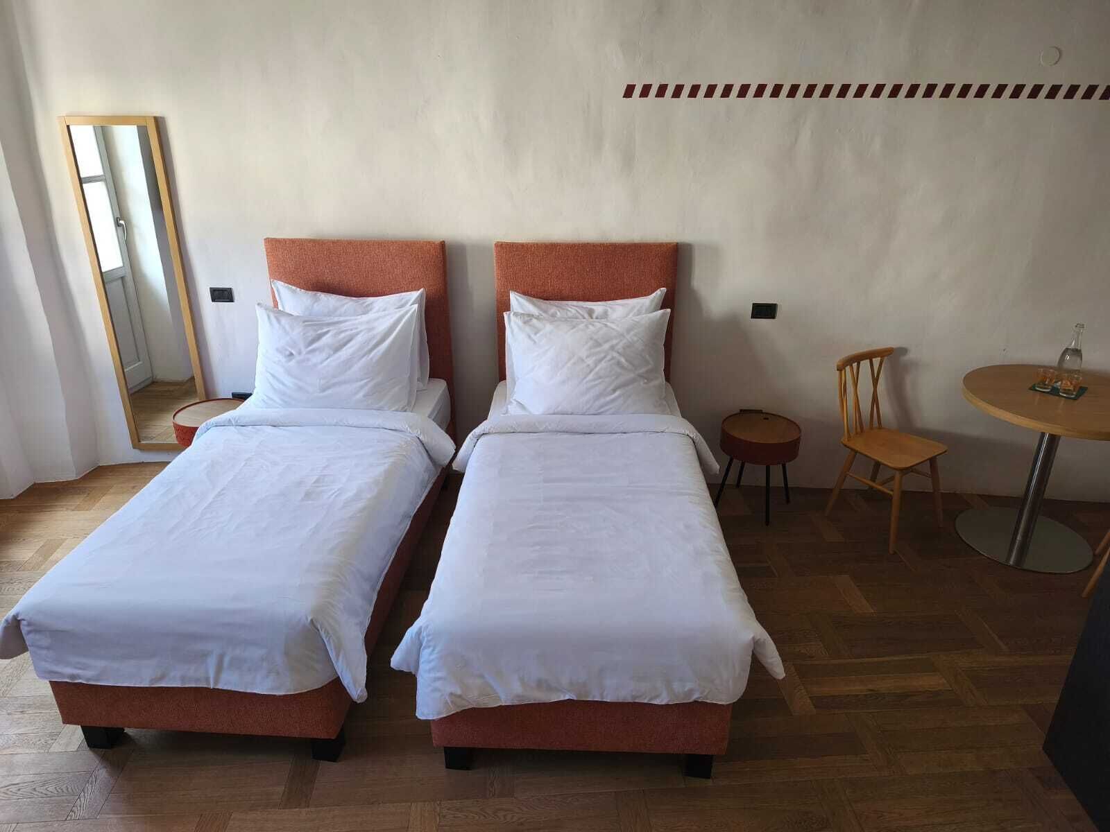 Quarto com duas camas de solteiro, uma mesa redonda com cadeiras, um espelho e uma pequena mesa redonda com garrafa e copos.