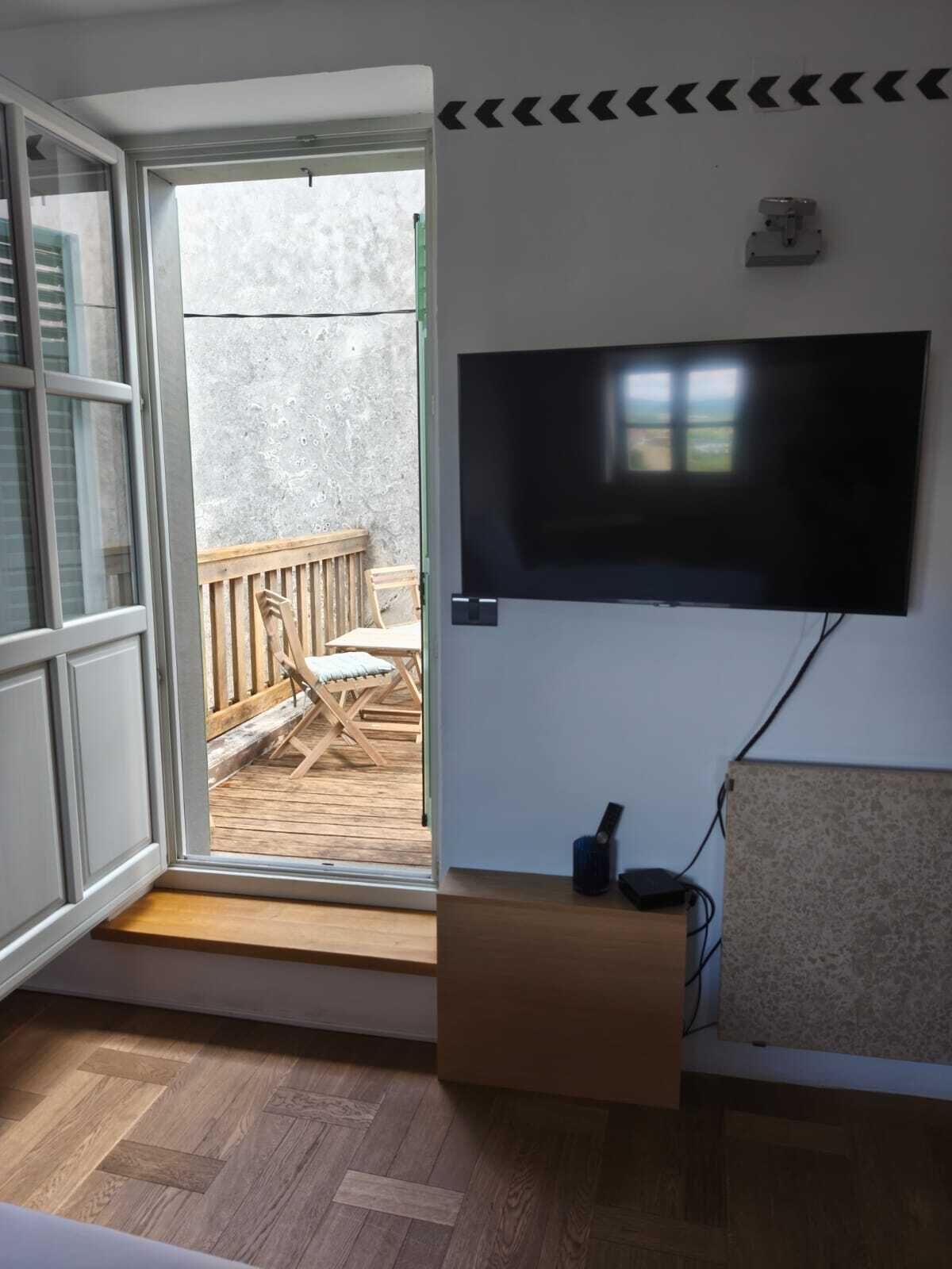 A room with a television mounted on the wall, a wooden bench, and a view of an outdoor deck with chairs.