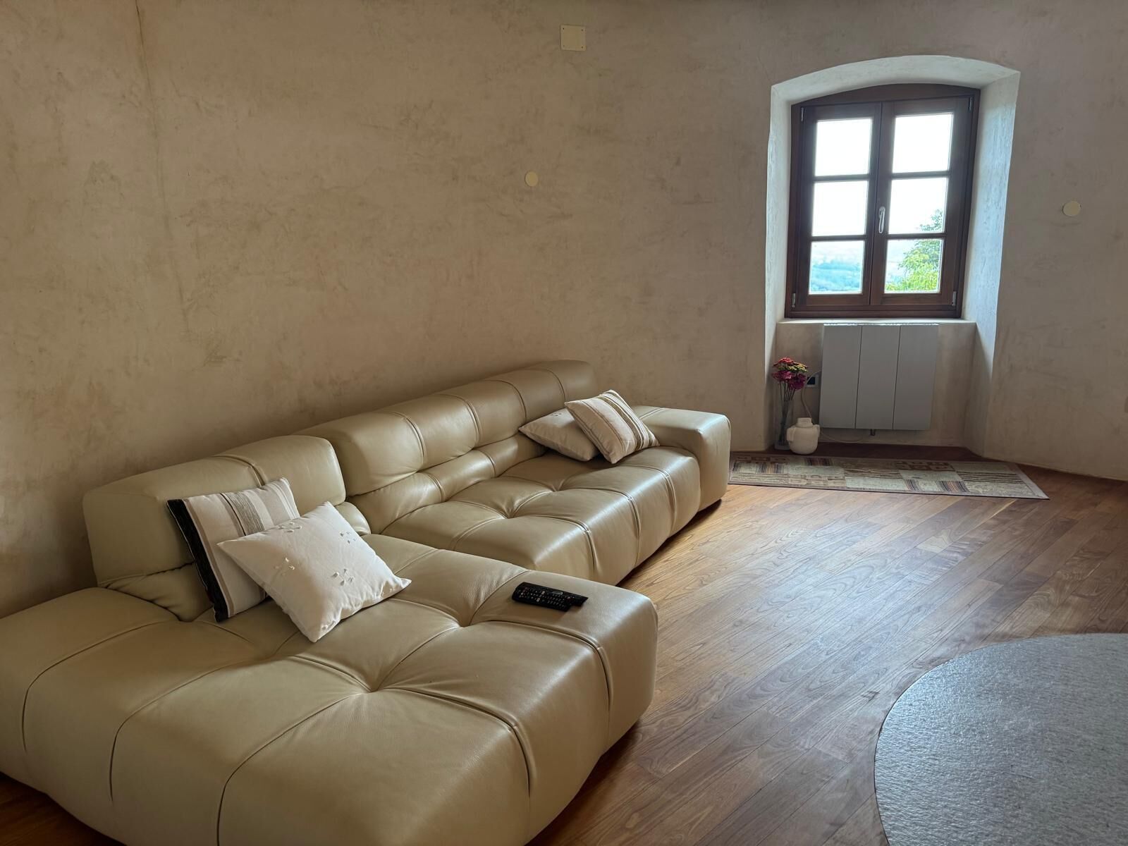 Ein beiges Ledersofa mit Kissen und einer Fernbedienung, ein Holzboden und ein Fenster mit Blick.