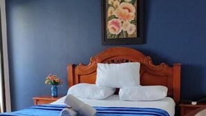 A neatly made bed with white pillows and a blue blanket, a wooden headboard, a small bedside table with a vase of flowers, and a framed painting on the wall. - Hotel Dorado Baq (Barranquilla)