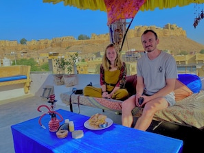 Een paar zit op een bed met uitzicht op de stad, een tafeltje met een shisha en lekkernijen, en een kamerplant.