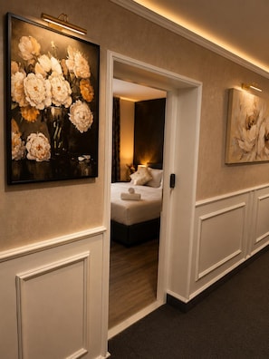 A hallway with a painting of flowers on the left, a bed with a white headboard in the background, and a door leading to another room. - Alsago Lux (Sesvete)