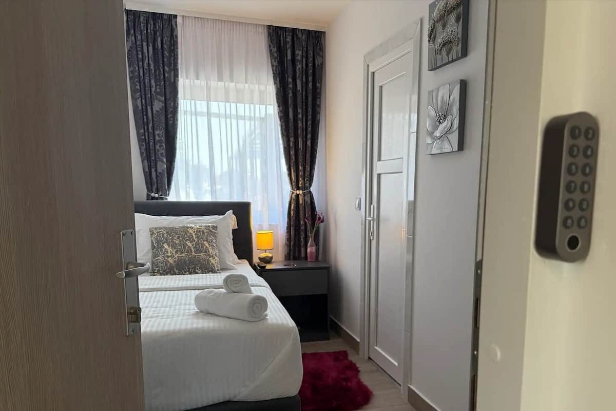 A hotel room with a bed, a nightstand, a lamp, a door, and a wall-mounted control panel.