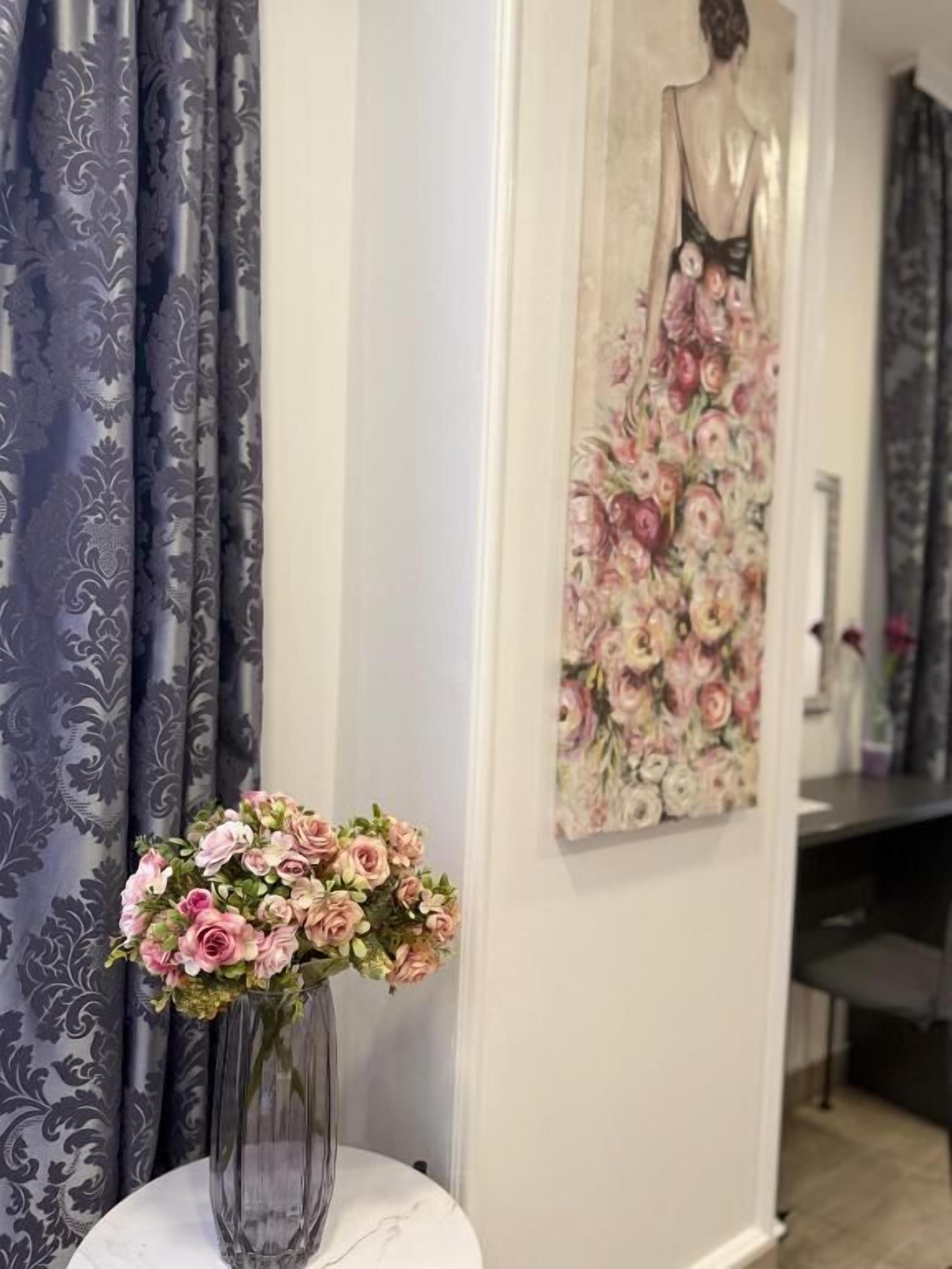 Una habitación con un jarrón de flores rosadas sobre una mesa blanca, un cuadro de un vestido en la pared y cortinas con estampado.