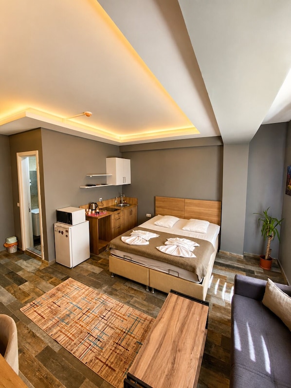 A compact hotel room with a bed, a small kitchenette, a seating area, and a bathroom.