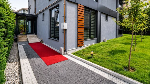 Un edificio moderno con alfombra roja que conduce a la entrada, un pequeño jardín y cielo despejado.