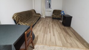 Un salón pequeño y escasamente amoblado, con un sofá, una silla, una mesita y un ventilador.