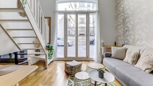 Un salon moderne avec un escalier, un canapé, une table basse et une vue sur la rue, visible à travers une porte vitrée.