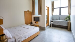 A hotel room with a bed, a TV, a small table, a bench, and a window with curtains. - The Quarters Business Hotel (Bangalore)