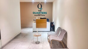 A reception area with a wooden reception desk, a computer, and a wheelchair. There are two chairs and a small table with a magazine. The walls are plain, and the floor is tiled. - The Quarters Business Hotel (Bangalore)