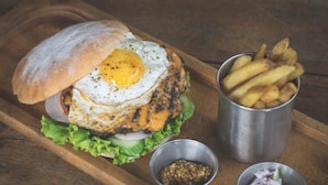 A burger with a fried egg, fries, and two condiments on a wooden tray. - The Quarters Business Hotel (Bangalore)