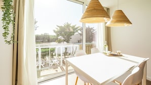Una habitación luminosa con una mesa y sillas blancas, ventanas amplias y lámparas de madera colgantes.