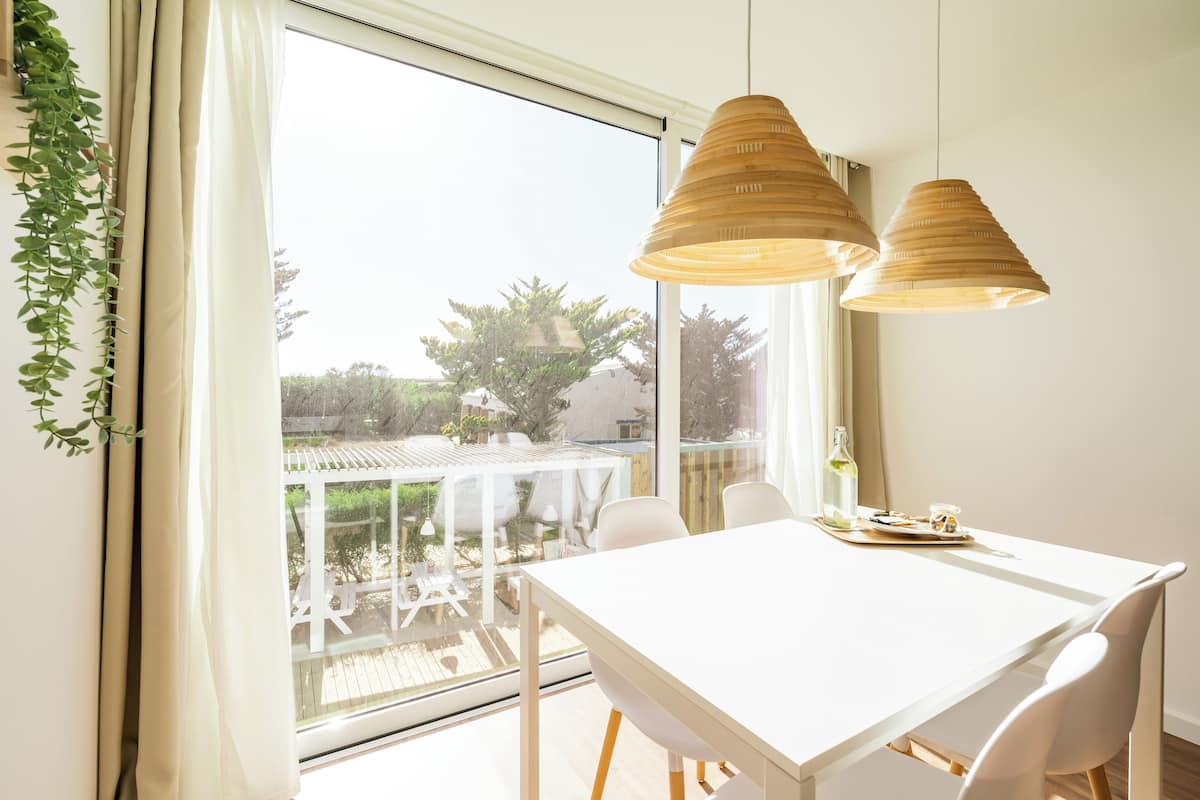 Une pièce lumineuse avec une table et des chaises blanches, de grandes fenêtres et des lampes en bois suspendues au plafond.