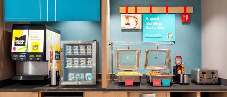 A breakfast station with a coffee machine, a refrigerator with beverages, and labeled food containers.