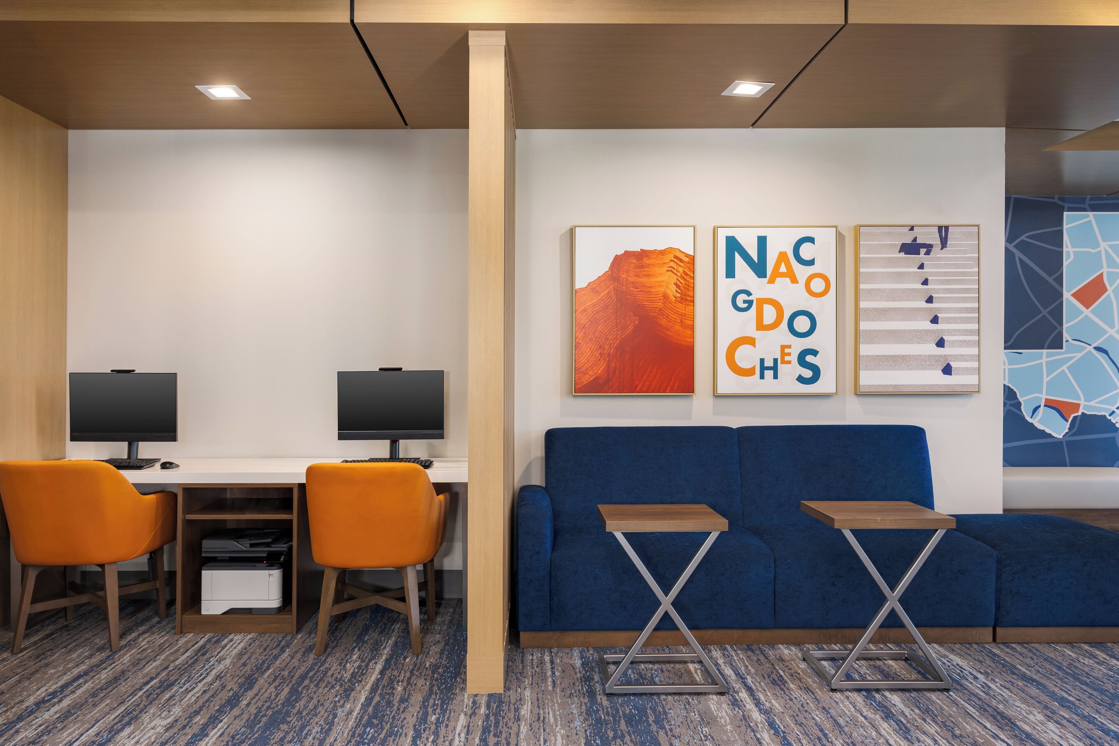 A modern office space with a blue sofa, two orange chairs, a desk with two monitors, and a small table.