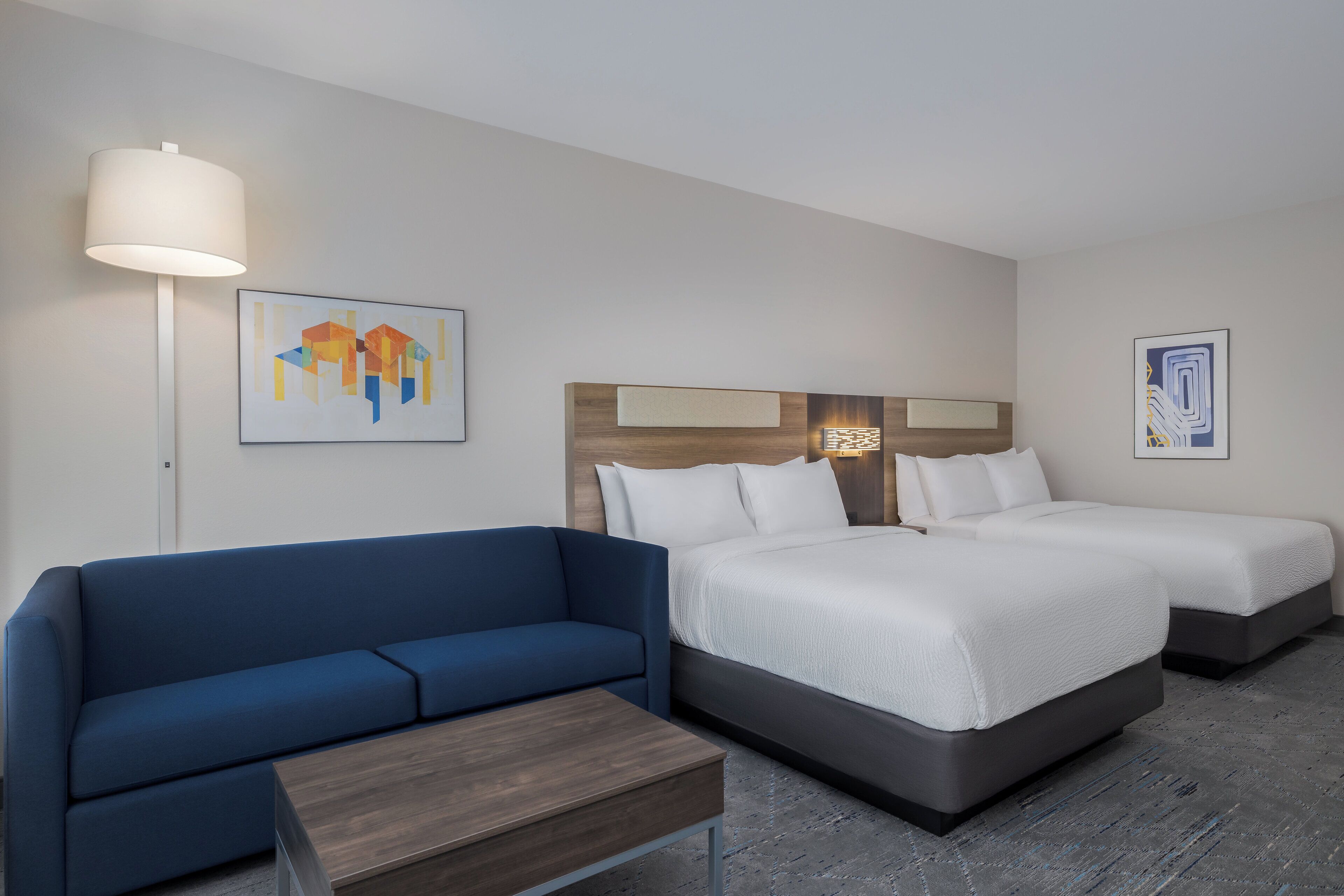 A hotel room with a blue sofa, two single beds, a wooden headboard, and a floor lamp.