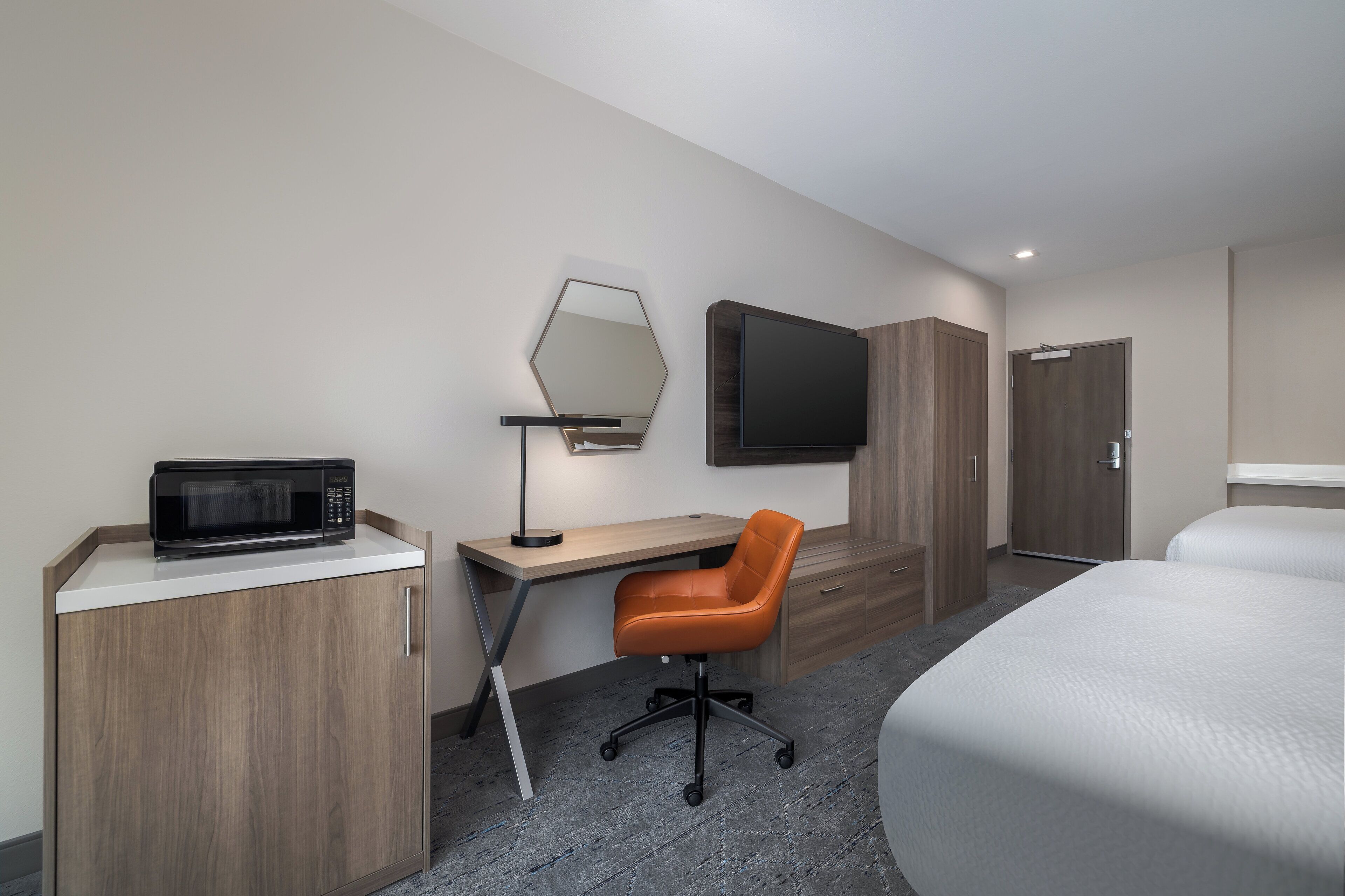 Una habitación de hotel moderna con un escritorio de madera, un microondas negro, una silla de oficina naranja, una cama grande y un televisor de pantalla plana montado en la pared.