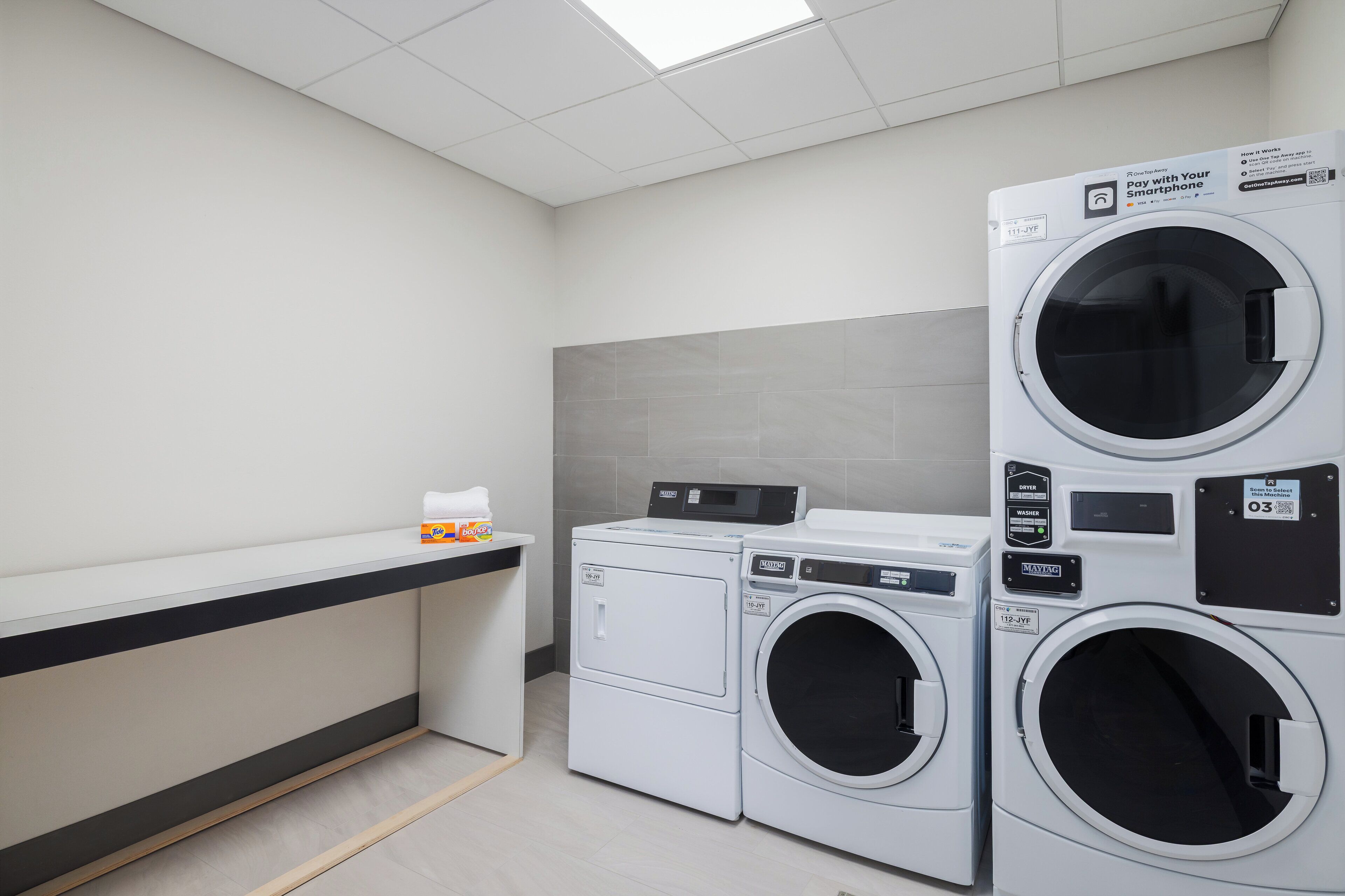 A laundry room with stacked washers and dryers, a counter with a towel dispenser, and a grey tiled wall.
