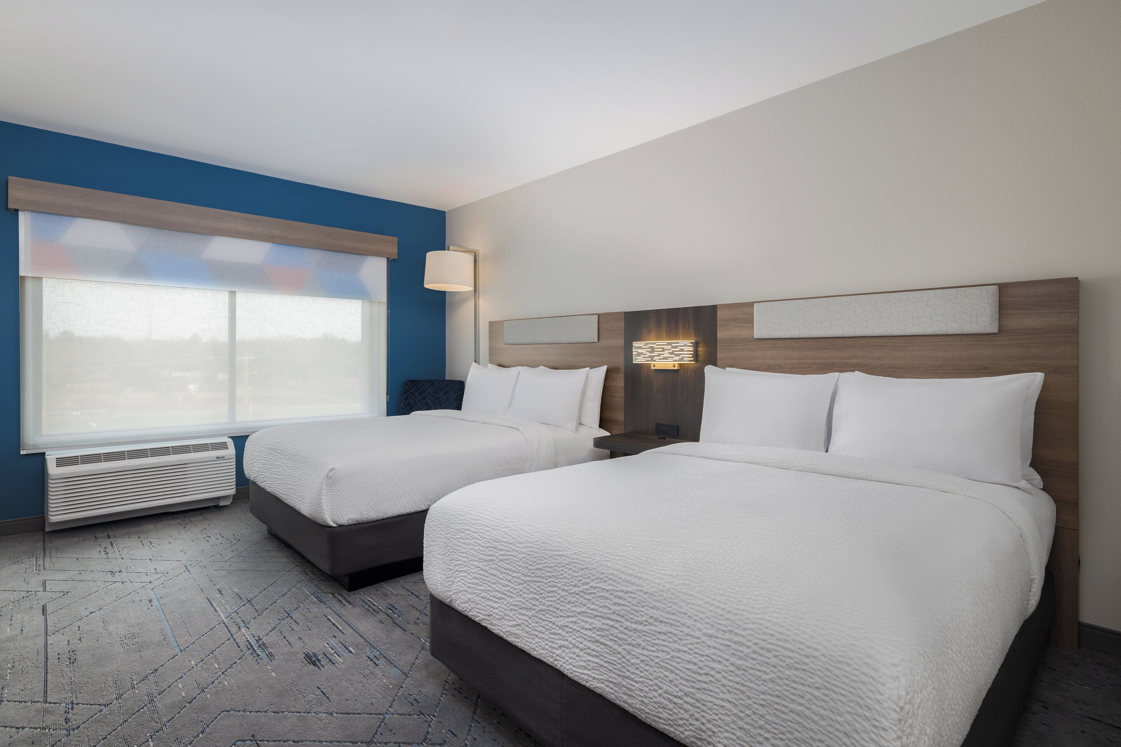 Habitación de hotel con dos camas, un ventanal grande con persianas, un calentador en la pared y una mesita de noche con lámpara.