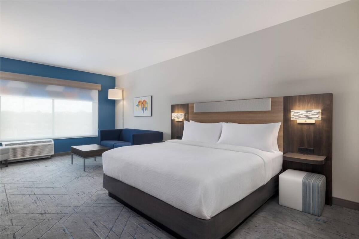 Ett modernt hotellrum med en stor säng, en blå accentvägg, en liten sittgrupp och ett fönster med persienner.
