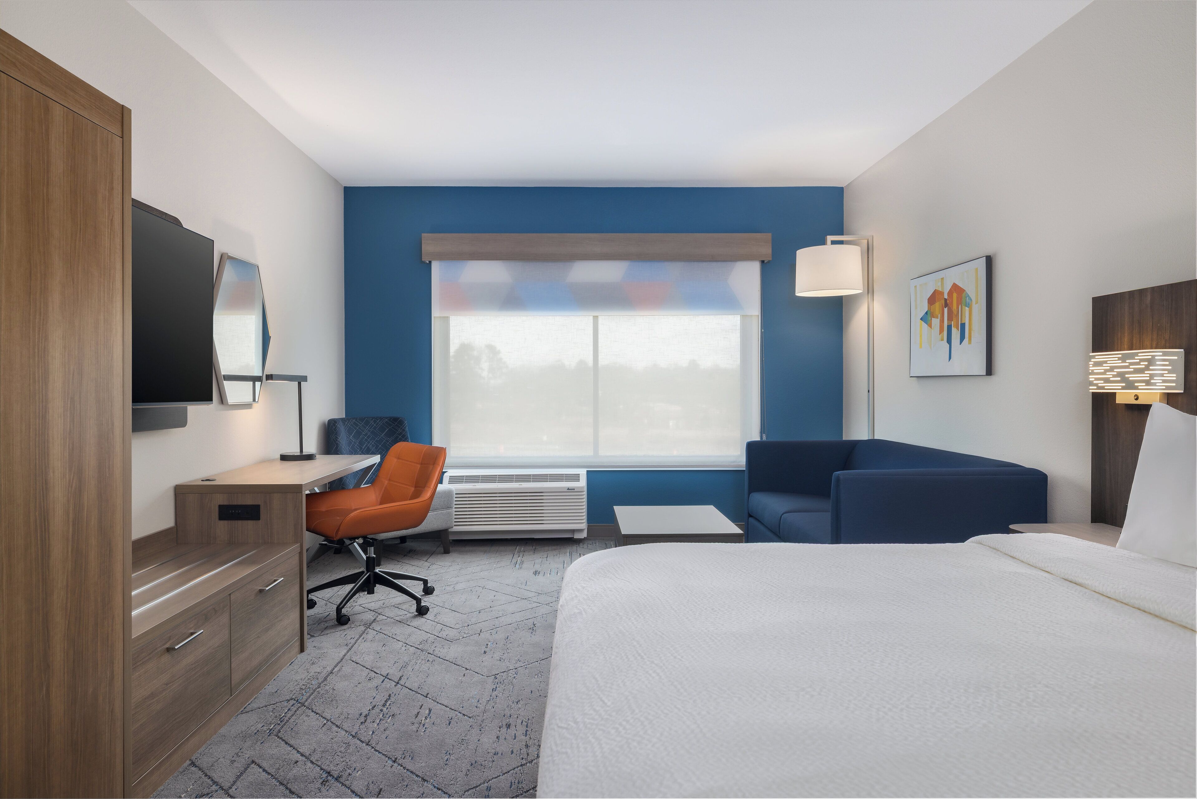 Habitación de hotel con cama, escritorio con silla, sofá, televisor y ventana con persianas.