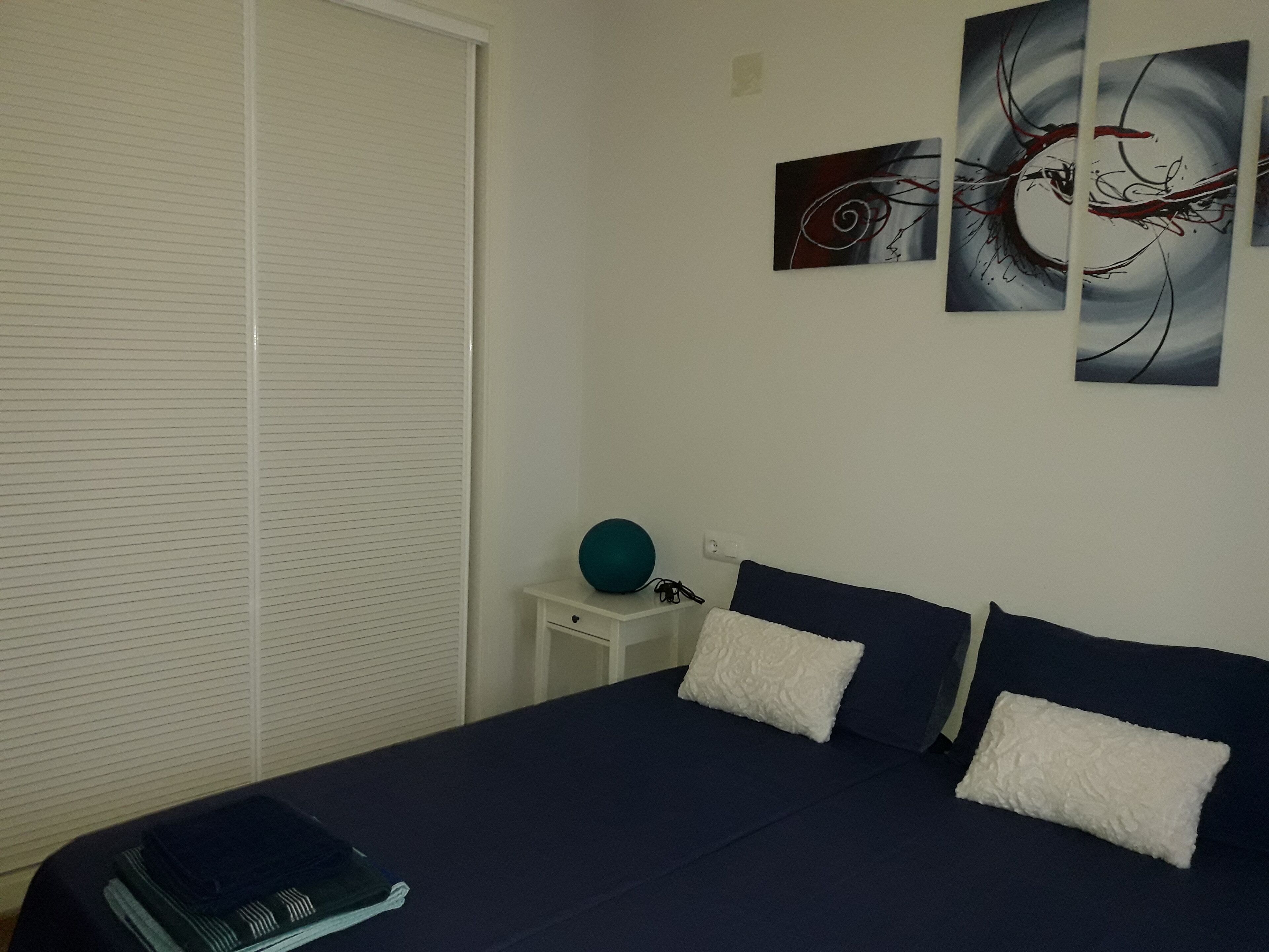 Een slaapkamer met een groot bed, twee kussens, een nachtkastje met een blauwgroen voorwerp en abstracte wanddecoratie.