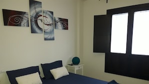 Ein Schlafzimmer mit Bett, Kissen, einem Nachttisch, einem Fenster und abstrakem Wandbild.
