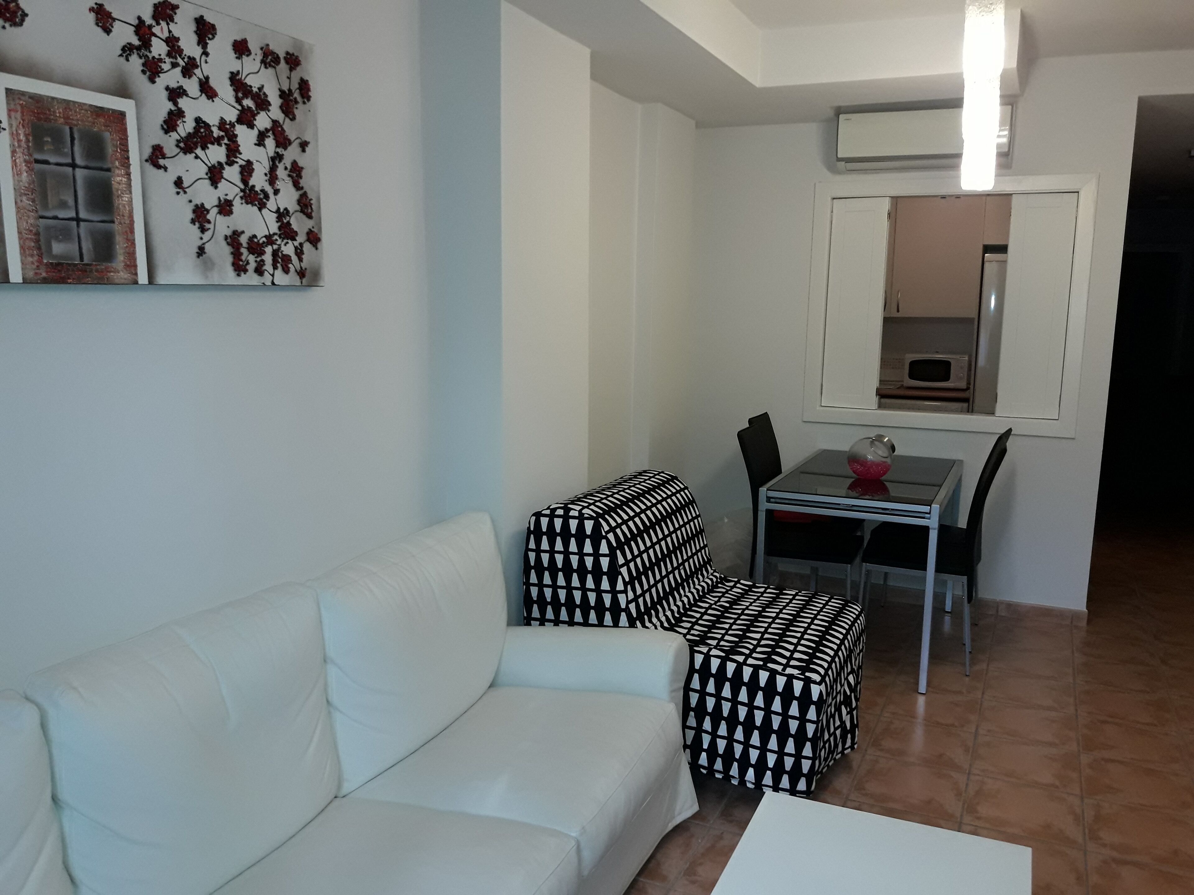 A living room with a white sofa, a patterned ottoman, a glass coffee table, and a dining area with a white table and chairs.