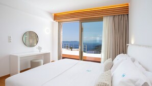 Une chambre d’hôtel avec un grand lit, un miroir rond, un bureau blanc et une vue sur la mer, visible à travers une porte-fenêtre coulissante.