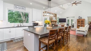 A modern kitchen with white cabinets, a large island with bar stools, a dining area with a table and chairs, and a TV in the living room.