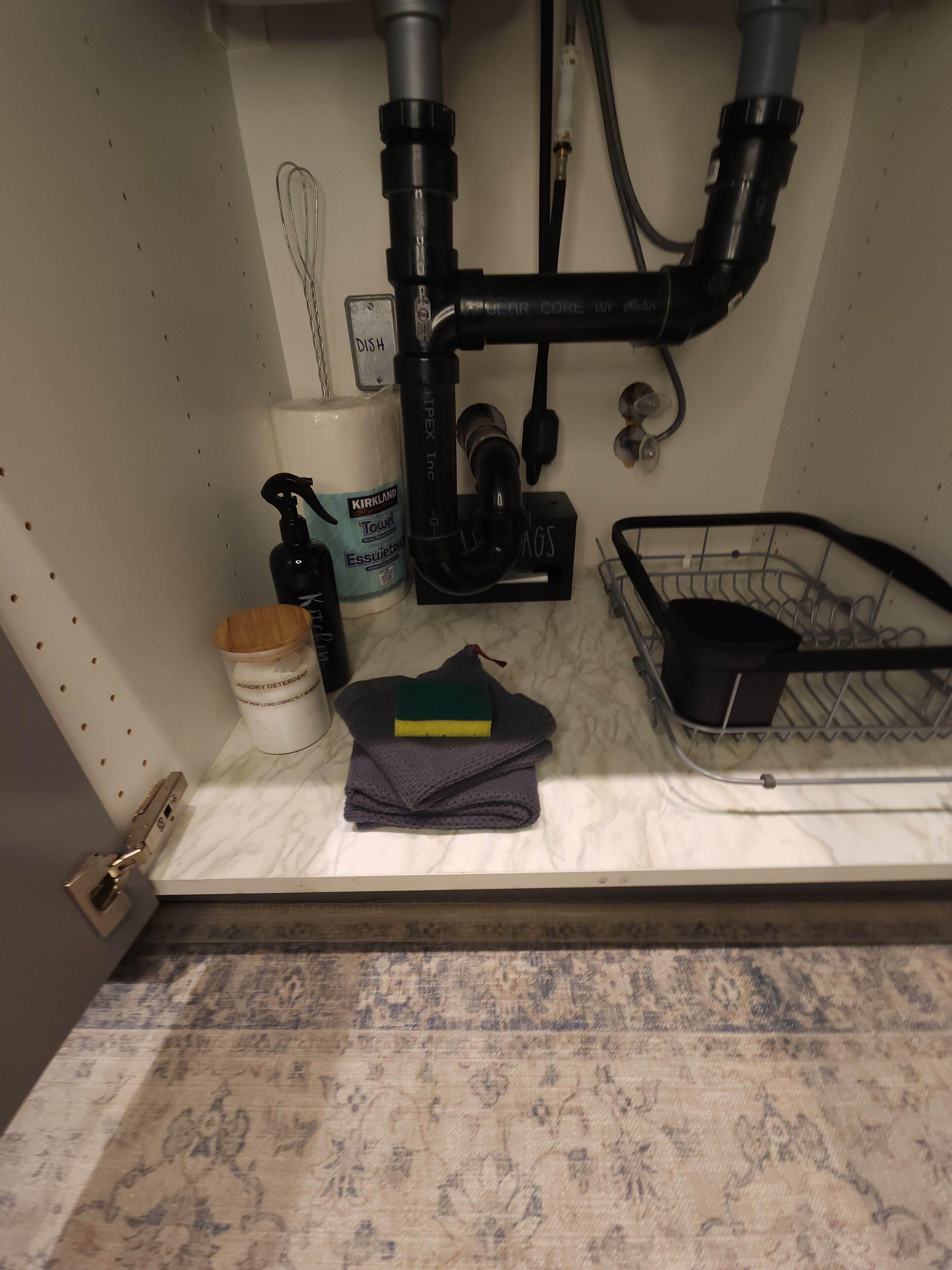 A kitchen sink with a dish rack, a towel dispenser, and a bottle of Kirkland Essentio detergent.
