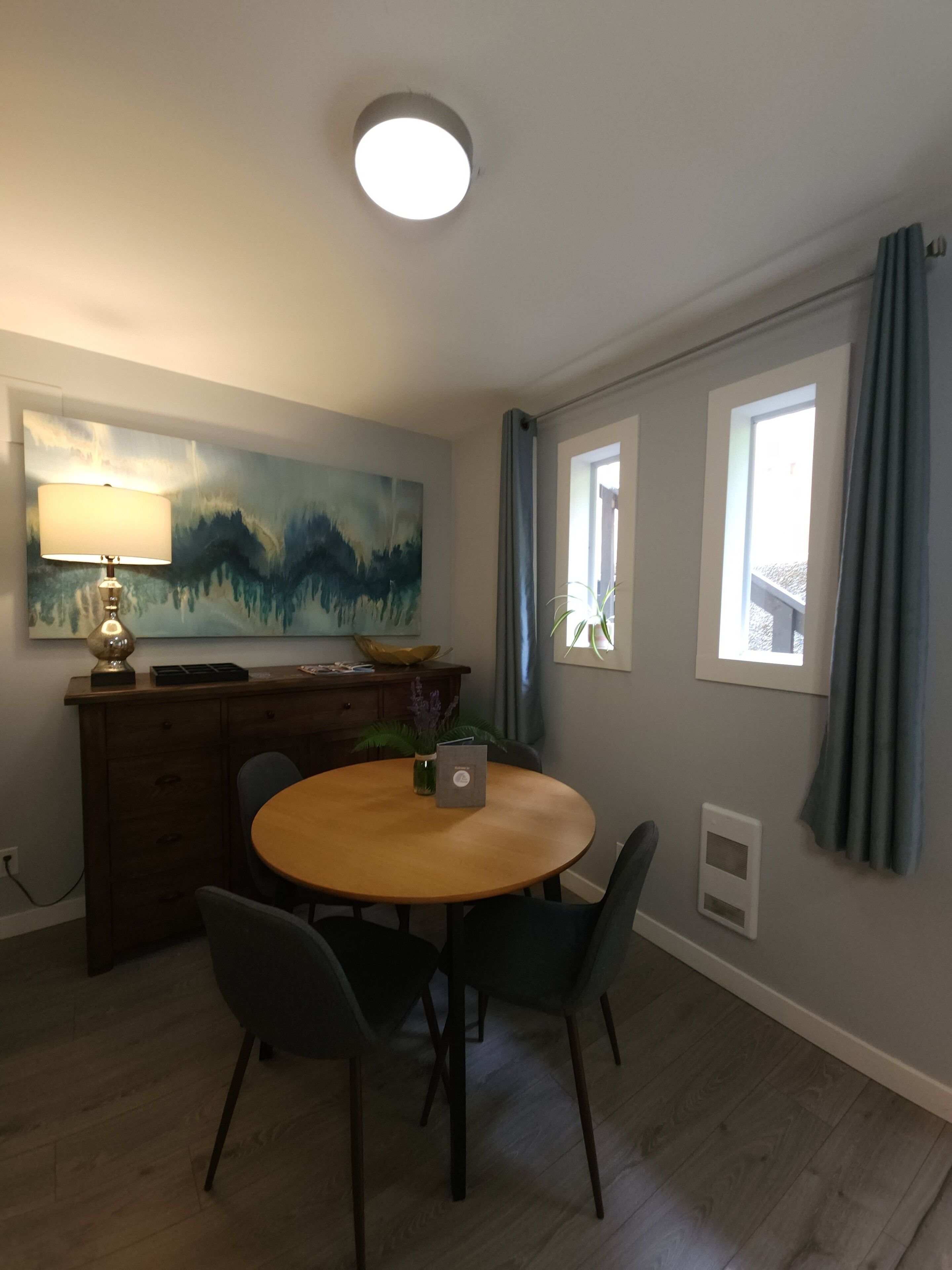 A dining area with a round table, chairs, a lamp, a painting, and a window with curtains.