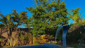 A swimming pool with a waterfall feature, surrounded by tropical vegetation and a clear blue sky. - SEASONAL RENTAL BEACH HOUSE ARACAJU SERGIPE CLOSED CONDO FRONT OF THE SEA (Mosqueiro Aracaju)