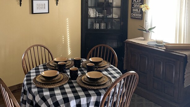 A dining room with a checkered tablecloth, wooden chairs, a black cabinet with glass doors, and a 'Welcome Home' sign on the wall.